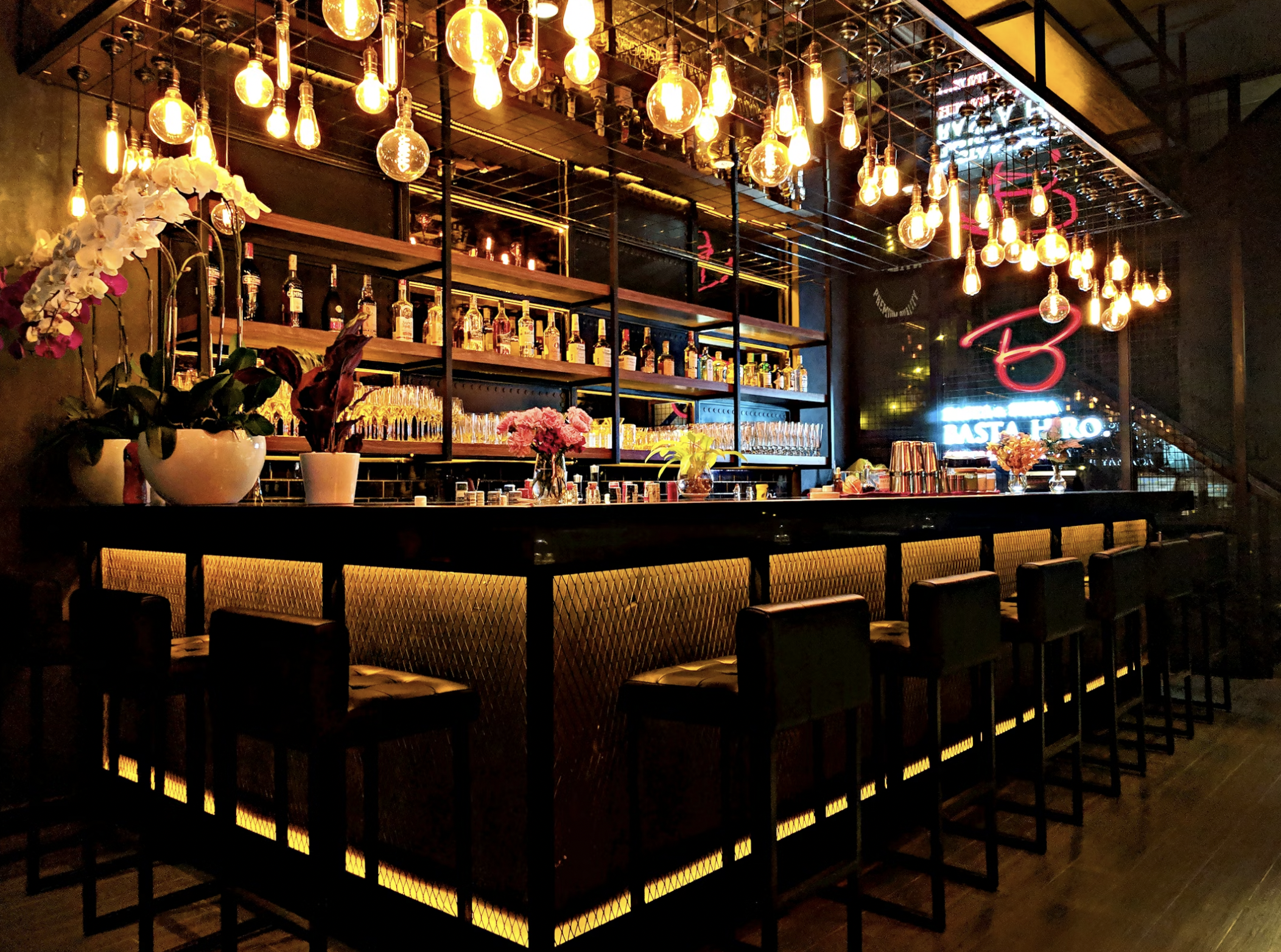 Dimly lit bar interior with warm hanging Edison bulbs, liquor shelves, and glassware along a backlit counter.