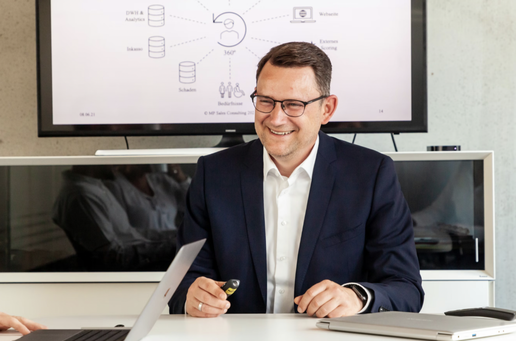 Smiling businessman in a navy suit sits at a table, holding a marker, with a diagram on a screen behind him.