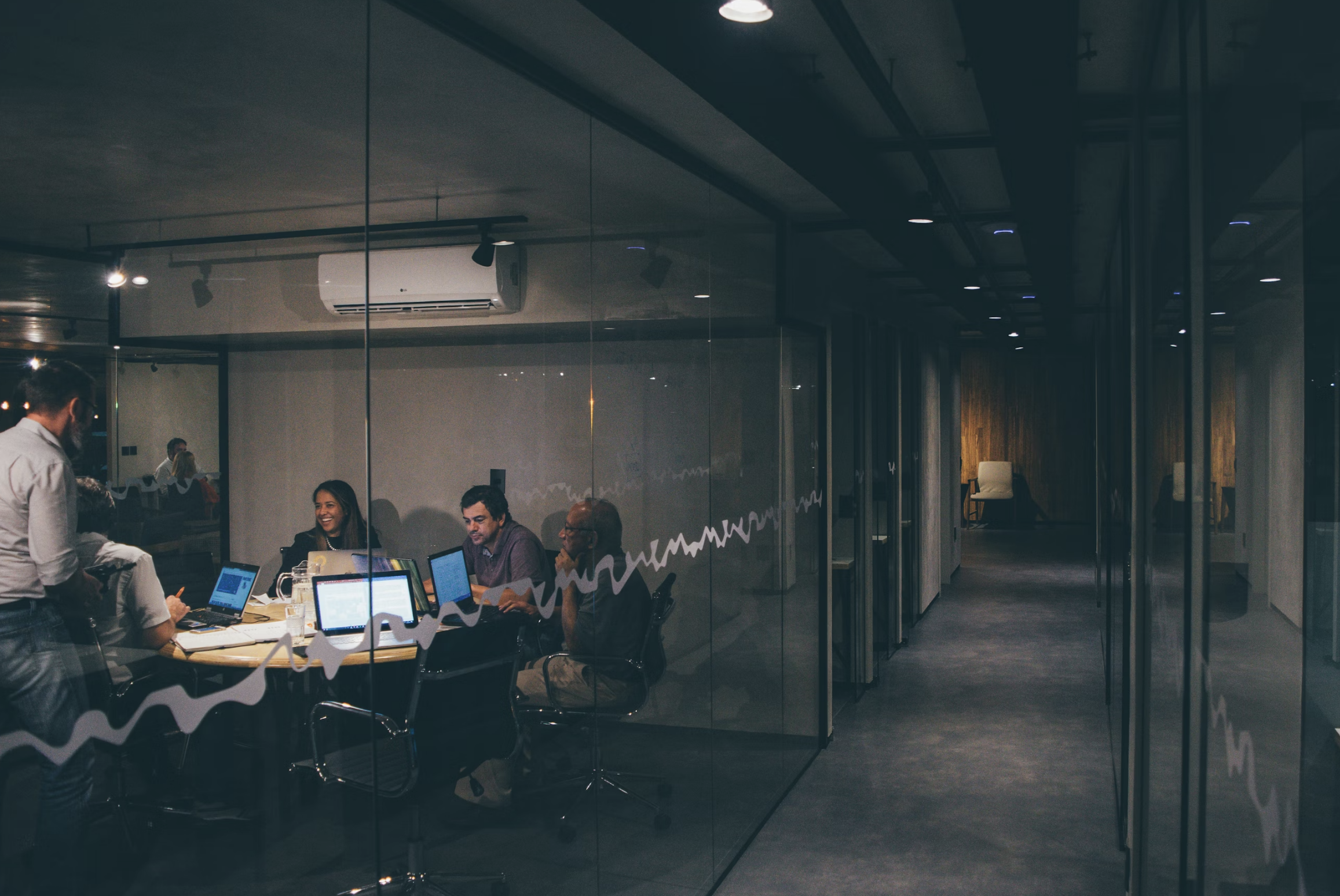Office conference room with glass walls; people seated around a table with laptops