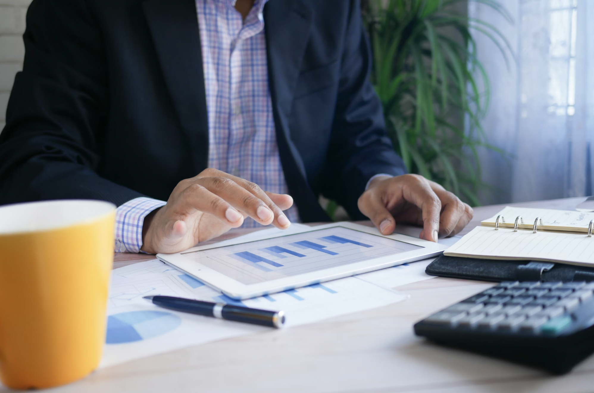 Person in a blazer reviews charts on a tablet at a desk; yellow mug in foreground.