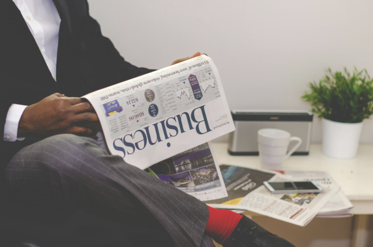 Person in a dark suit reading a newspaper at a desk in an office setting with a plant and coffee mug nearby.
