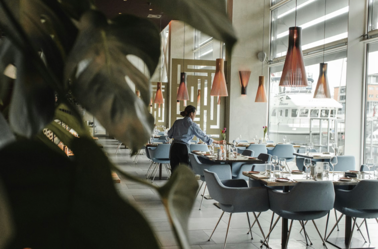 Restaurant dining area with blue chairs, orange pendant lights, and a server arranging a table near large windows overlooking a marina.
