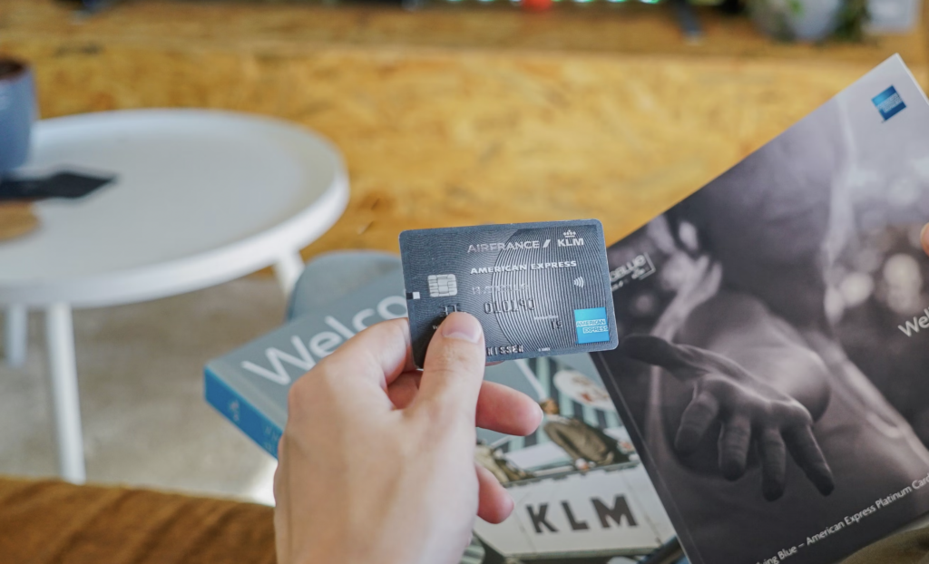Close-up of a hand holding a blue KLM American Express credit card, with a travel brochure and magazine in the blurred background.