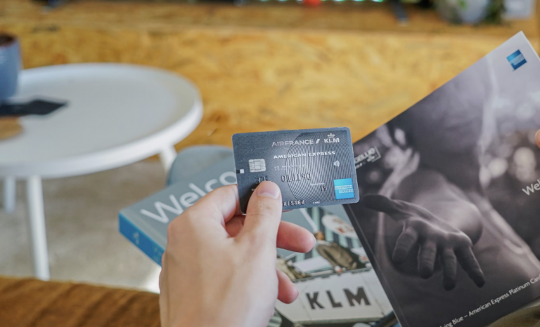 Close-up of a hand holding a blue KLM American Express credit card, with a travel brochure and magazine in the blurred background.