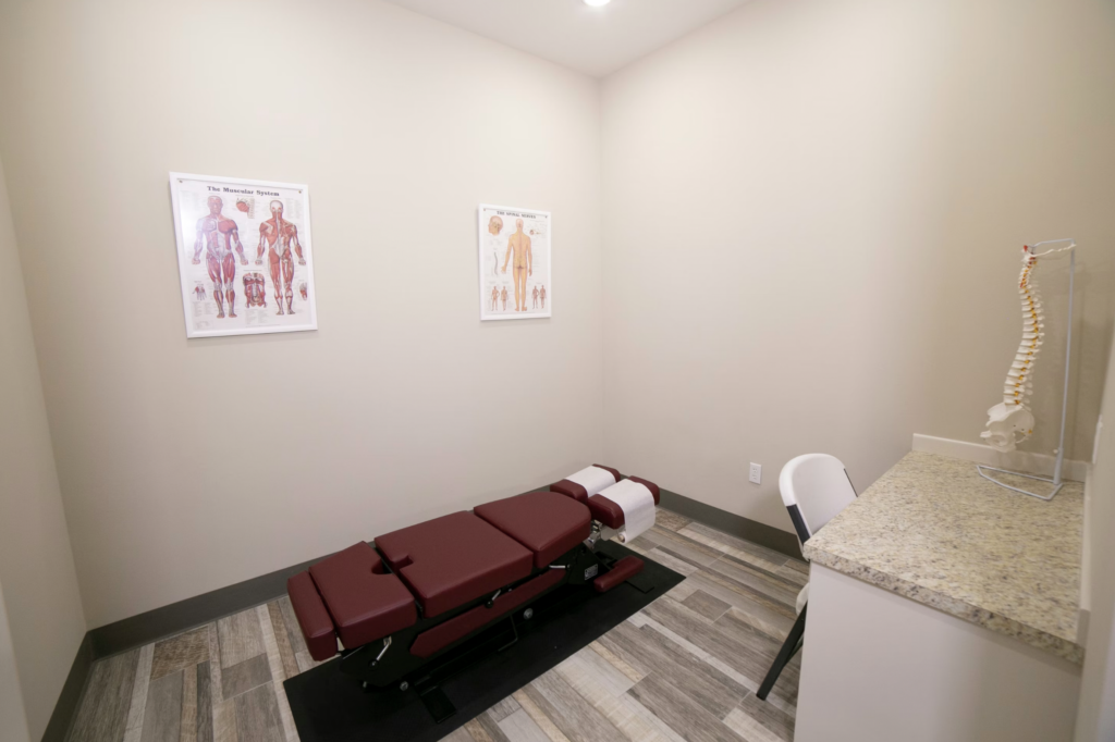 A treatment room with a red adjustable examination table, anatomy posters on the walls, and a spine model near a granite counter.