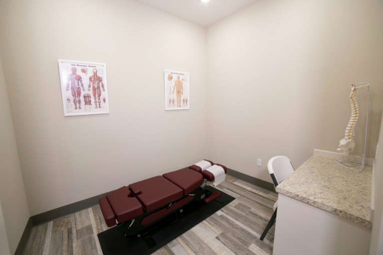 A treatment room with a red adjustable examination table, anatomy posters on the walls, and a spine model near a granite counter.
