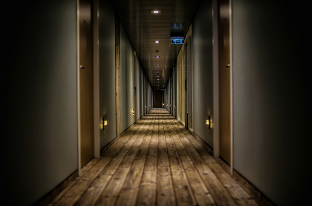 Dim hotel-style corridor with doors on both sides, warm recessed lights, and a wooden floor receding into the distance.