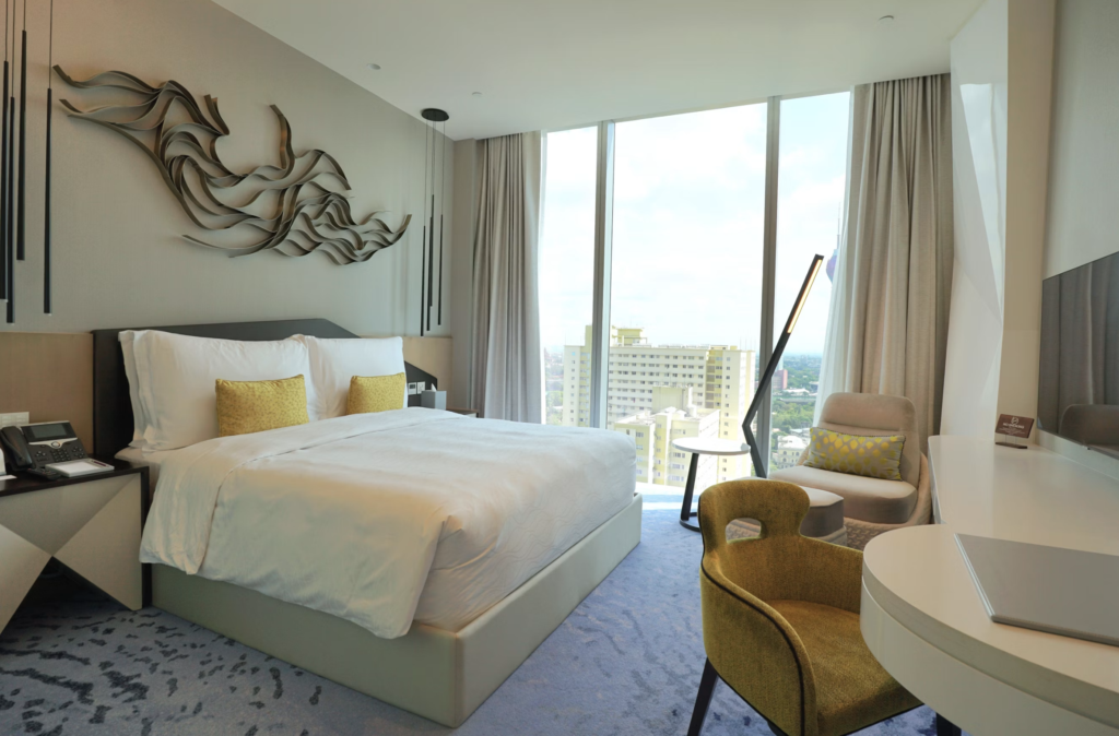 Hotel bedroom with a large bed, white linens, and two yellow accent pillows, a decorative wall sculpture above the headboard, and a seating area by floor-to-ceiling windows showing a cityscape. An desk and phone are at the bedside table.