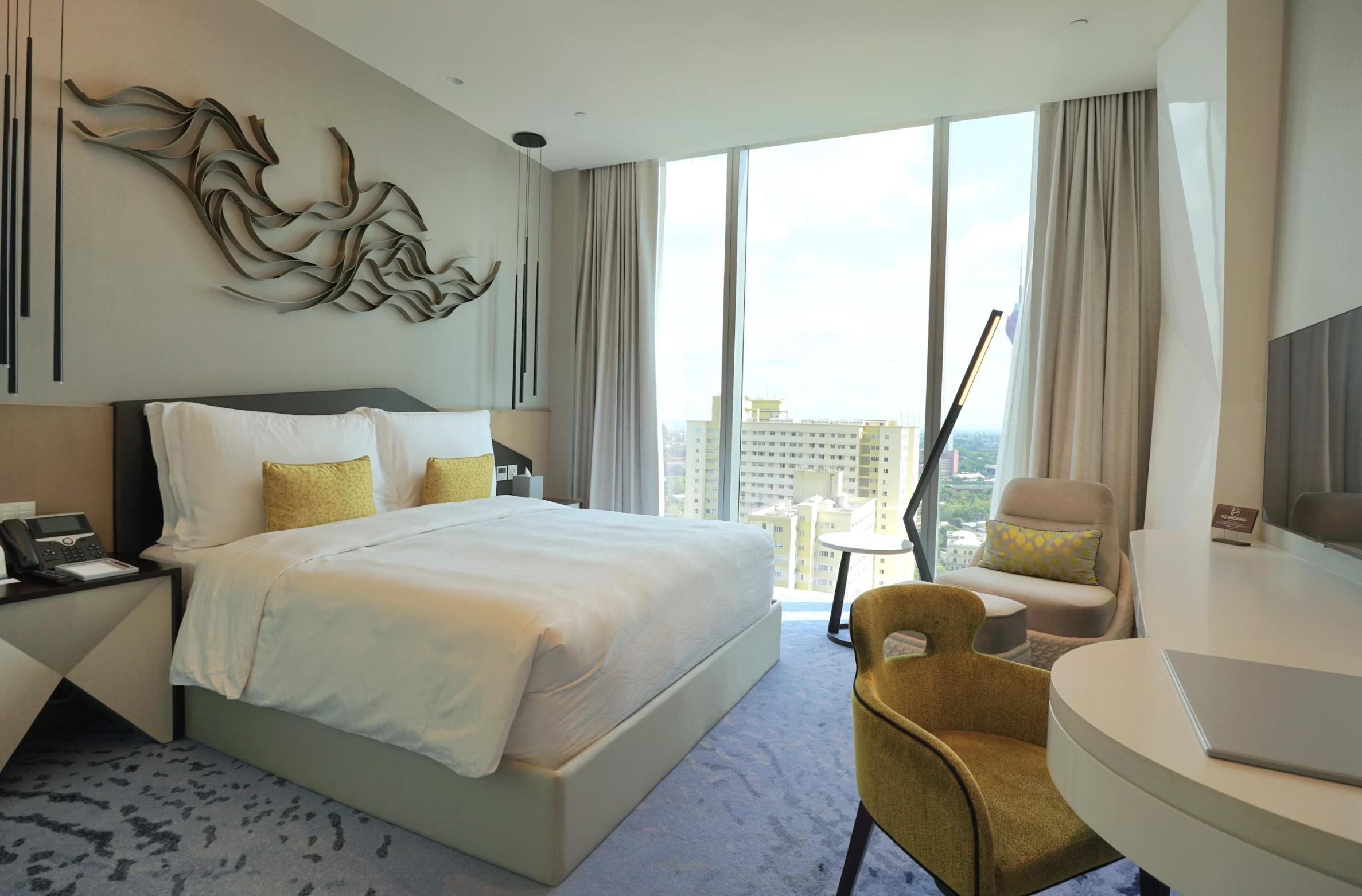 Hotel bedroom with a large bed, white linens, and two yellow accent pillows, a decorative wall sculpture above the headboard, and a seating area by floor-to-ceiling windows showing a cityscape. An desk and phone are at the bedside table.