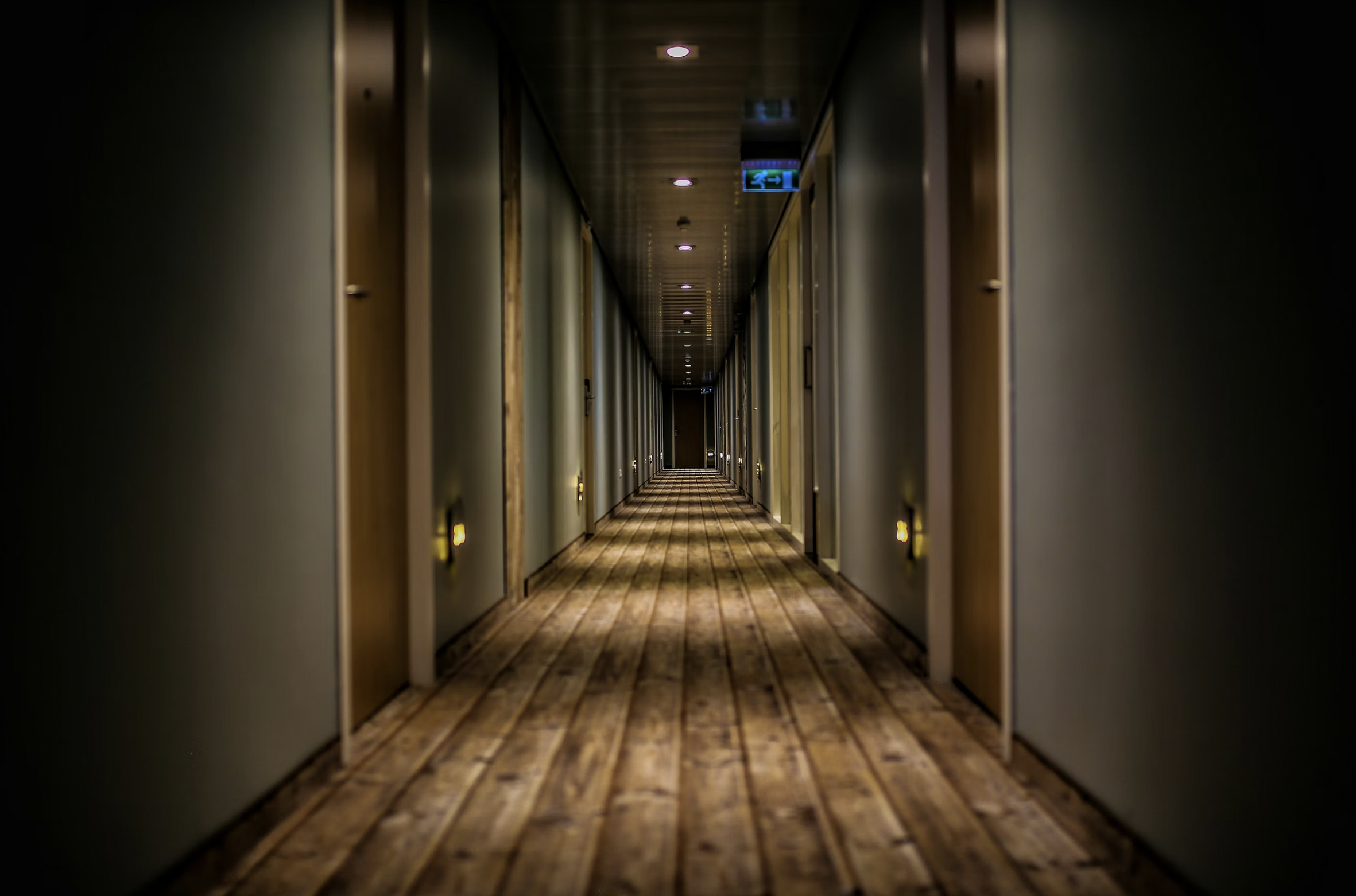 Dim hotel-style corridor with doors on both sides, warm recessed lights, and a wooden floor receding into the distance.