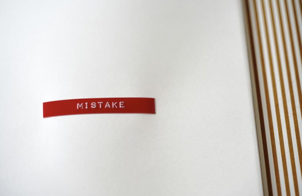 Red curved strip with the embossed text 'MISTAKE' on a white surface, with striped fabric visible on the right edge.