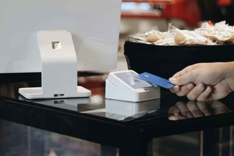 Customer paying with a blue card at a white card reader on a checkout counter; groceries in the background.