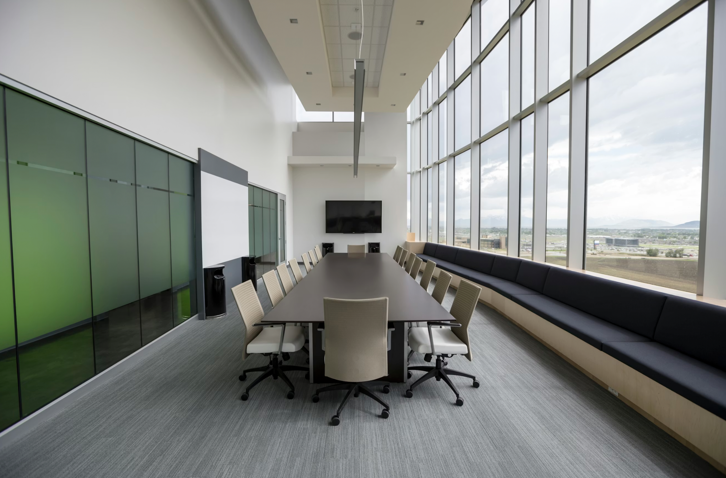 Modern conference room with a long dark table, beige rolling chairs, and a wall-mounted TV, beside large windows showing a cityscape.