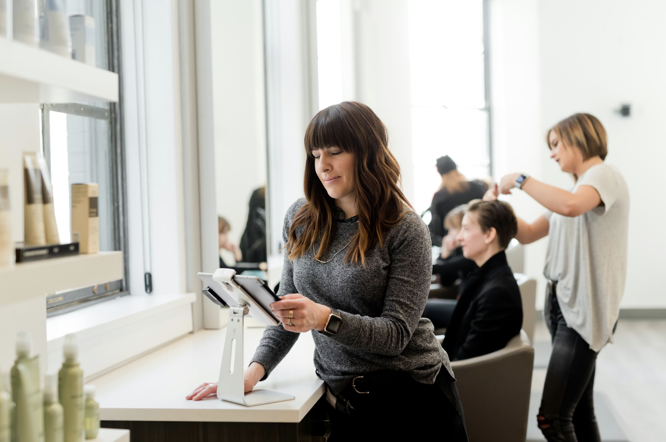 Woman at a salon station using a tablet on a stand, with hair products visible on the counter nearby, while another stylist works with a client in the background