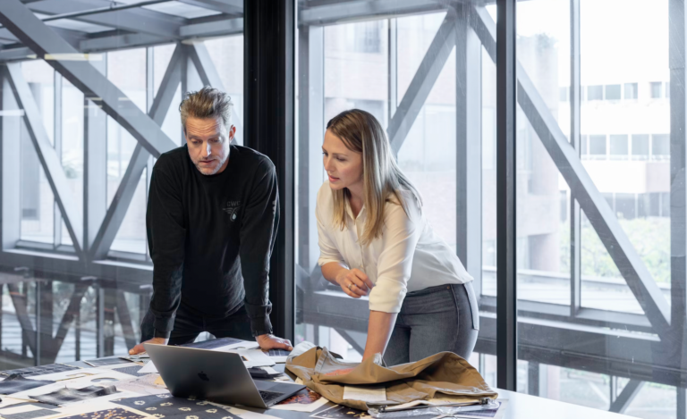 Two colleagues review documents on a glass conference table in a bright office, with a laptop and fabric swatches spread out before them.