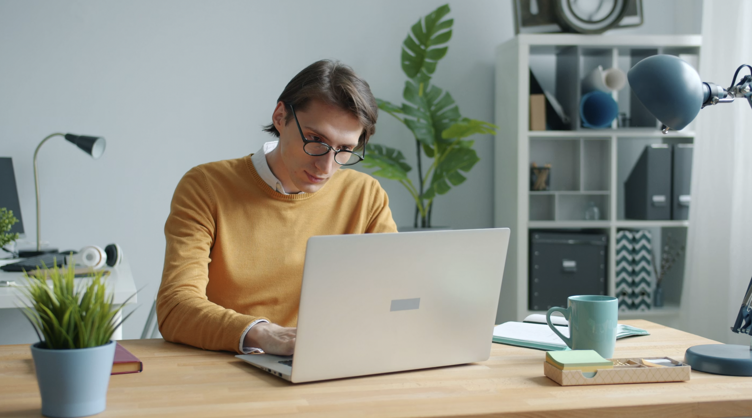 Person wearing an orange sweater and glasses types on a laptop at a tidy home office desk.