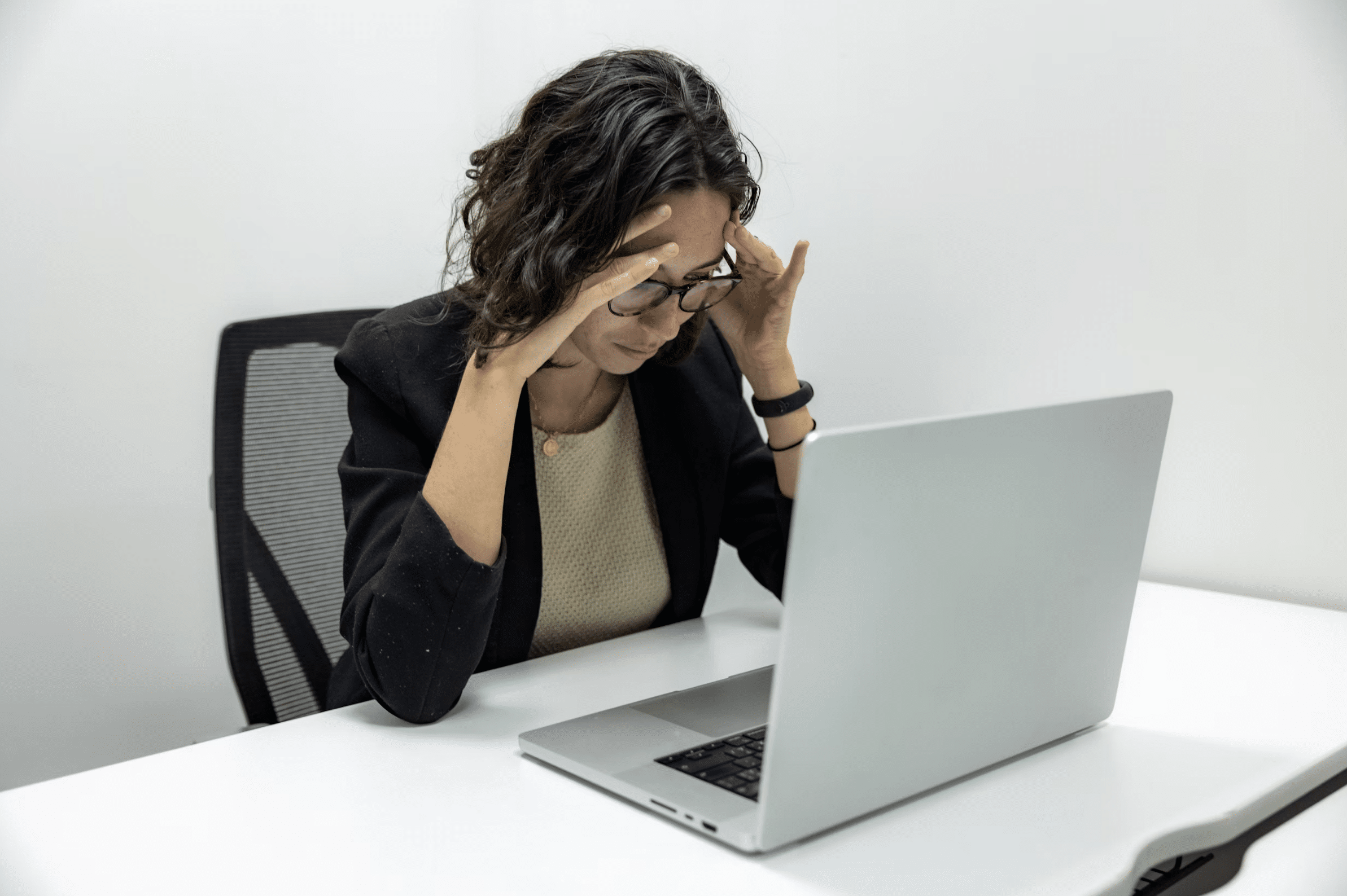 Professional woman at a white desk, hands on temples, looking stressed while working on a laptop . (office setting) [