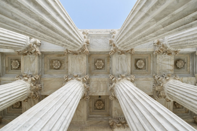 Low-angle view of marble columns with fluted shafts and ornate capitals against a blue sky, forming a grand architectural ceiling.Not necessary