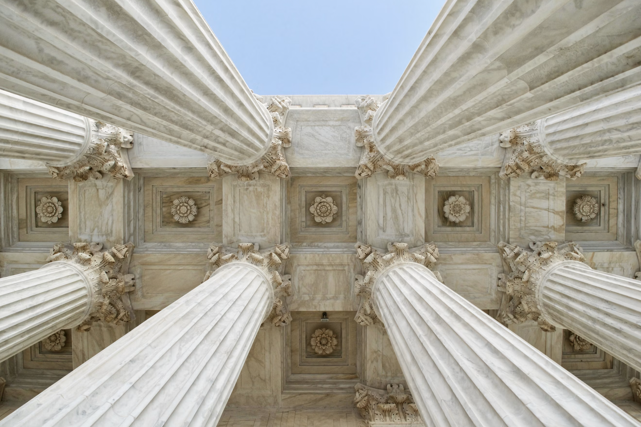 Low-angle view of marble columns with fluted shafts and ornate capitals against a blue sky, forming a grand architectural ceiling.Not necessary