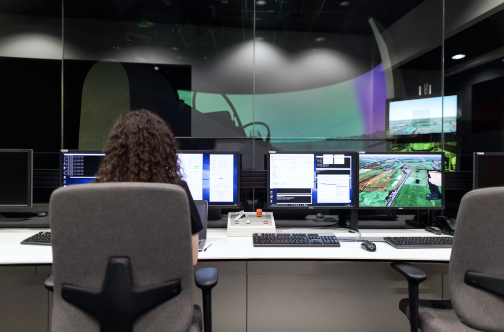 Back view of an operator with curly hair at a multi-monitor control desk in a dim operations room; glass wall with a colorful projection behind.
