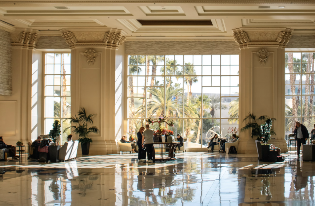 Bright hotel lobby with large windows and palm trees visible outside; guests sit at tables and plant pots are around.