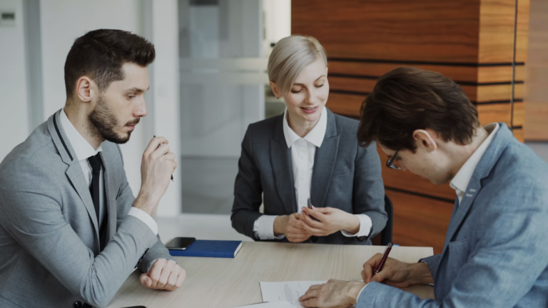 Three professionals in suits sit around a conference table reviewing documents in a modern office environment.