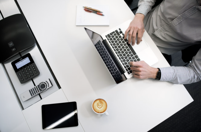 Top-down view of a person typing on a silver laptop at a white desk, with a notebook, pens, smartphone, tablet, and calculator nearby and a latte nearby.