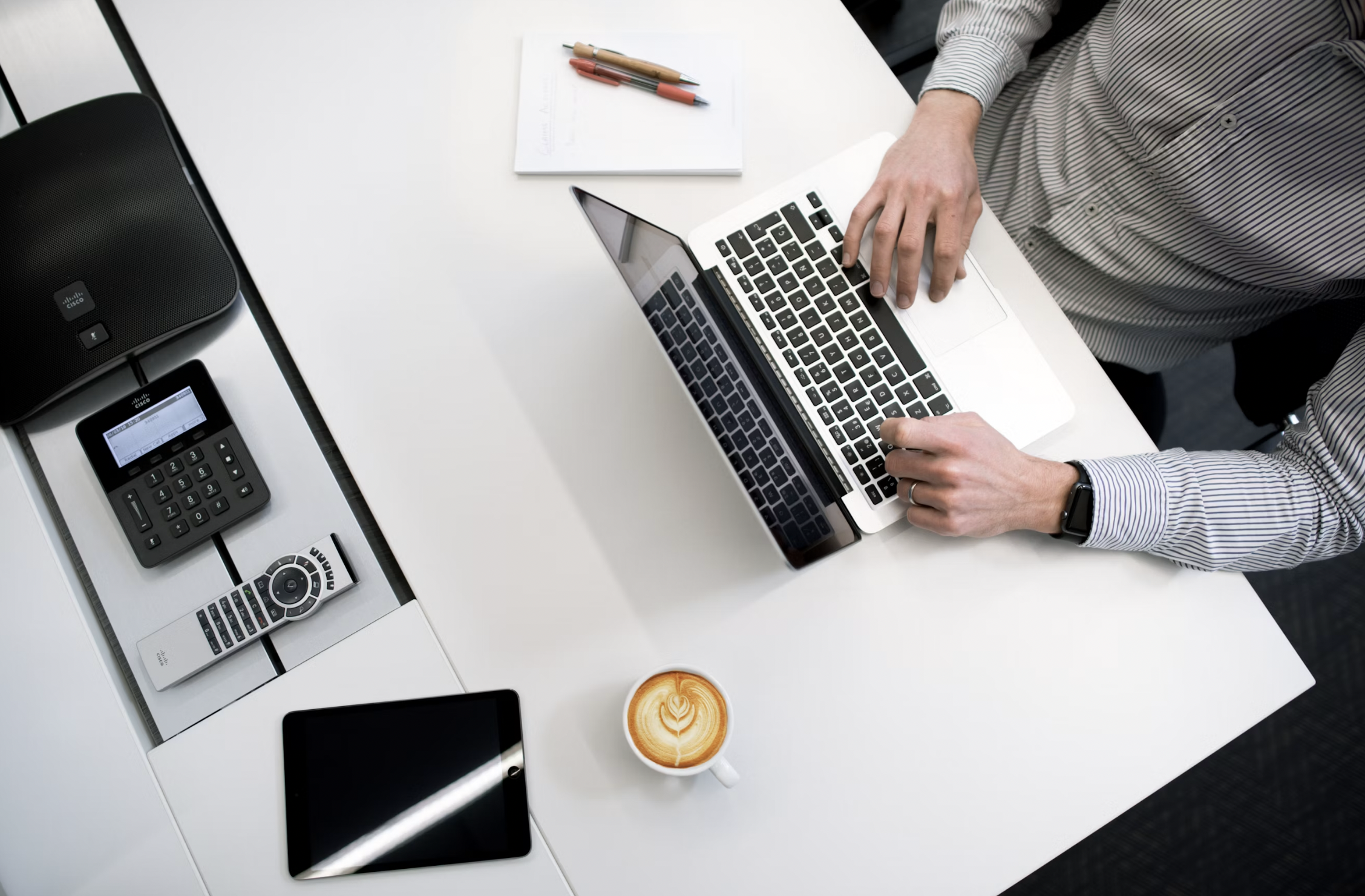Top-down view of a person typing on a silver laptop at a white desk, with a notebook, pens, smartphone, tablet, and calculator nearby and a latte nearby.