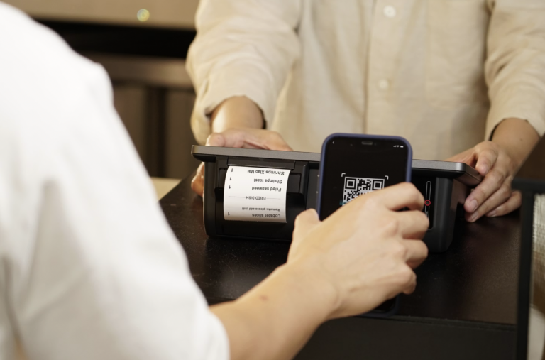 Hands at a payment terminal: smartphone near the reader and a printed receipt visible on the device.