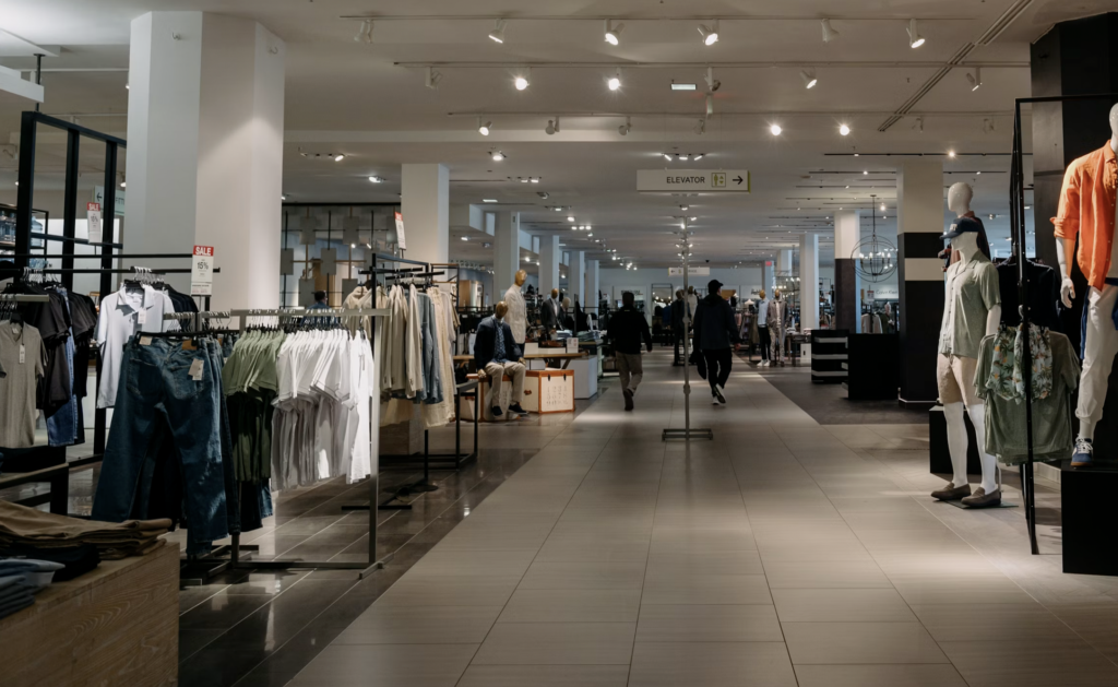 Spacious department store clothing area with racks of garments, mannequins on the right, and shoppers in the distance under bright track lighting.