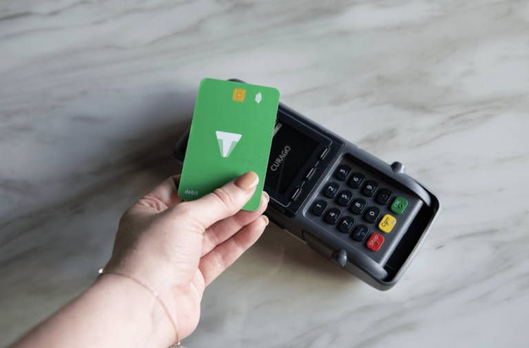 Hand holding a green debit card near a card payment terminal, about to swipe or insert the card for payment