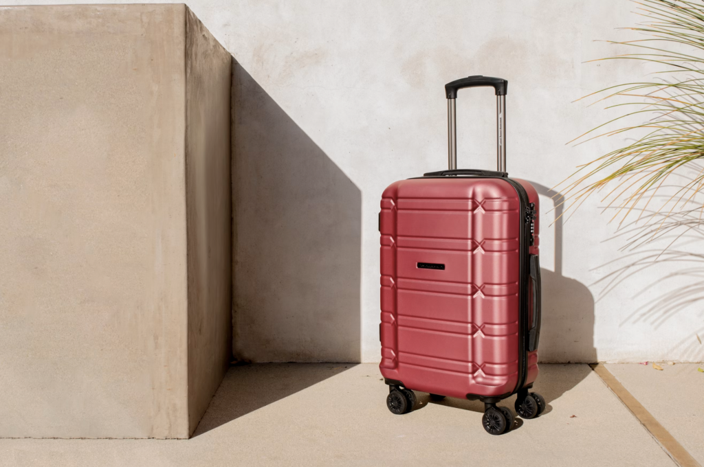 Red hard-shell suitcase with extended handle on a sidewalk beside a beige wall.