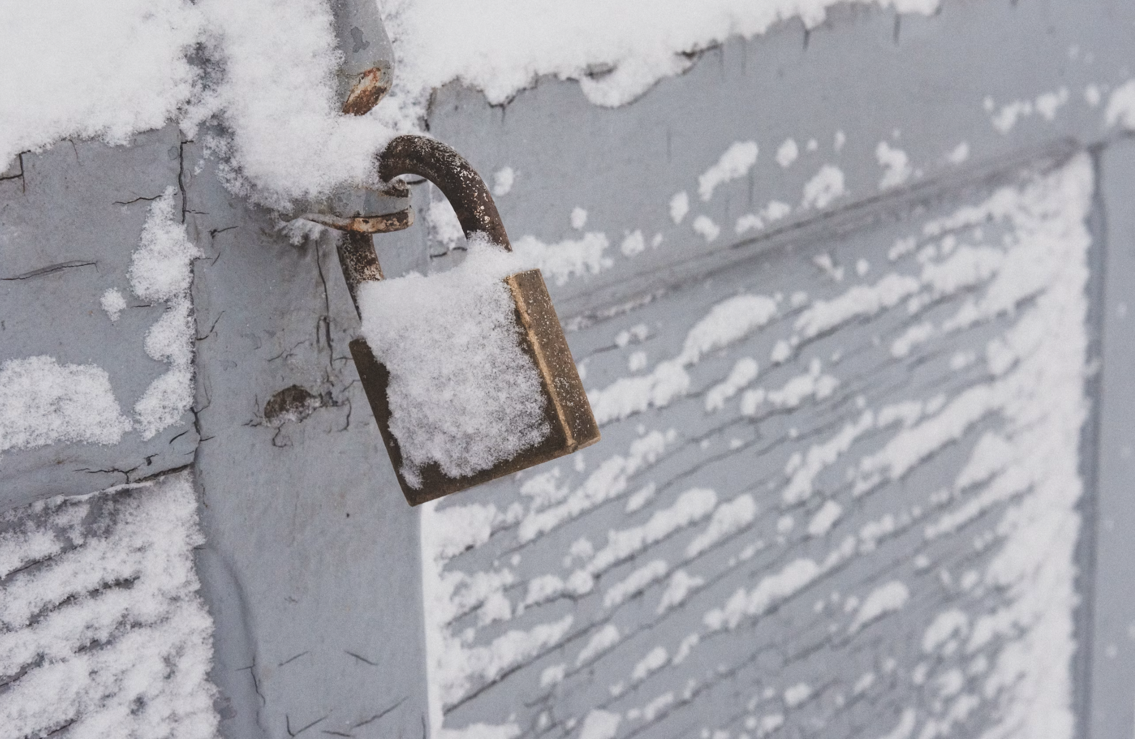 Rusty padlock on a weathered blue-gray door with powdery snow on the latch area outside.