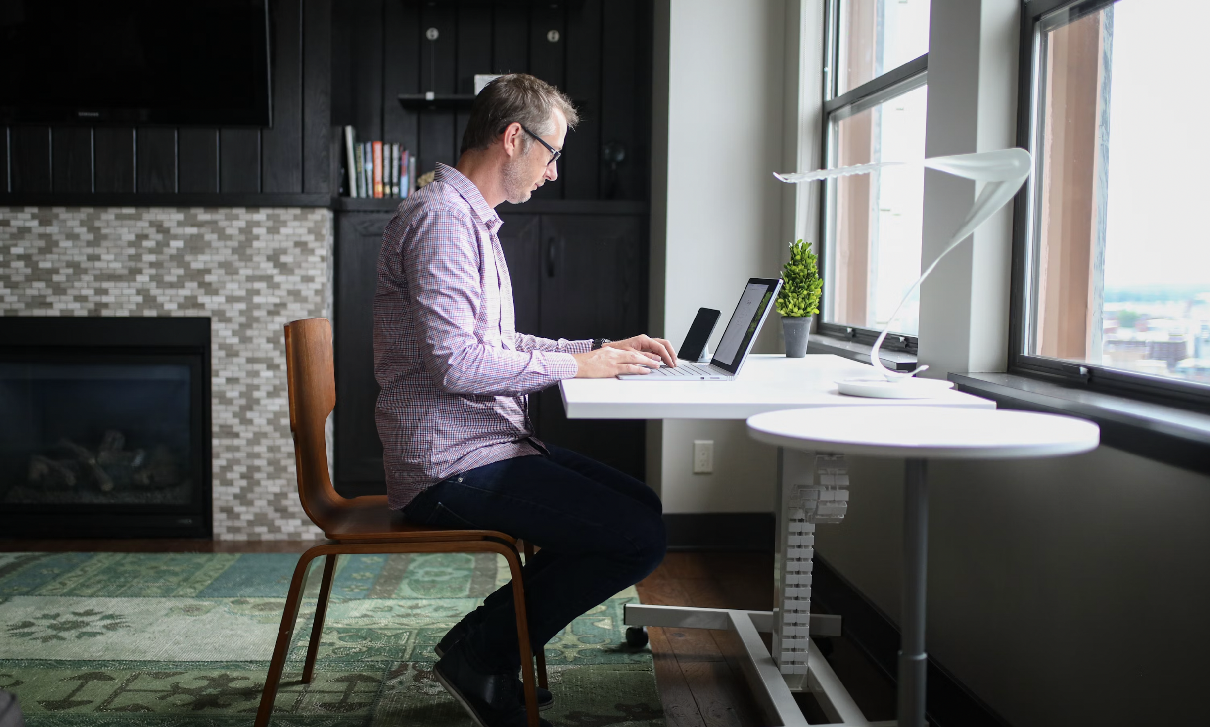Man in glasses typing on a laptop at a white desk by a large window with a small potted plant nearby