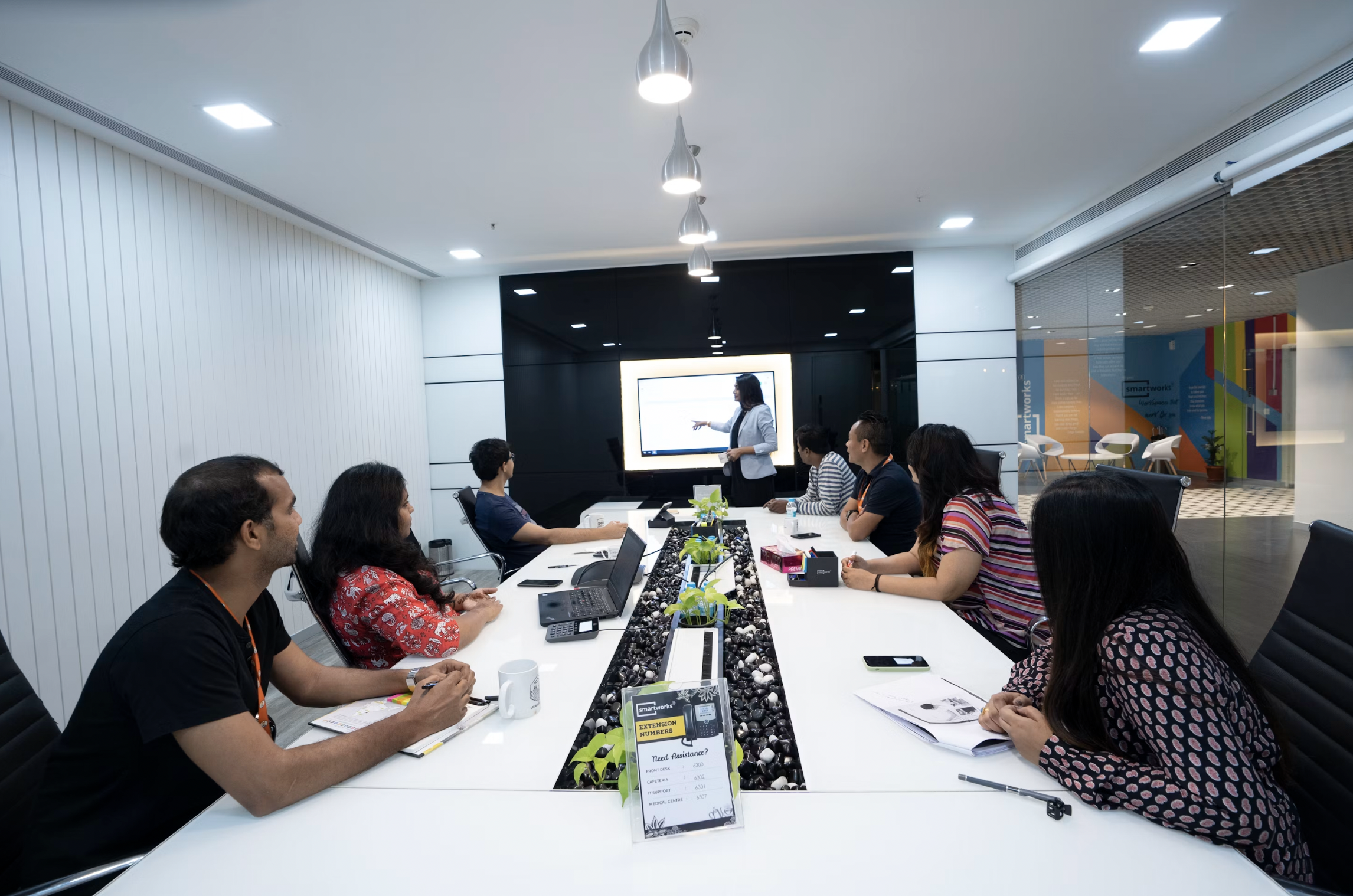 Group of professionals seated around a long white conference table watching a presenter at the front with a screen behind them.