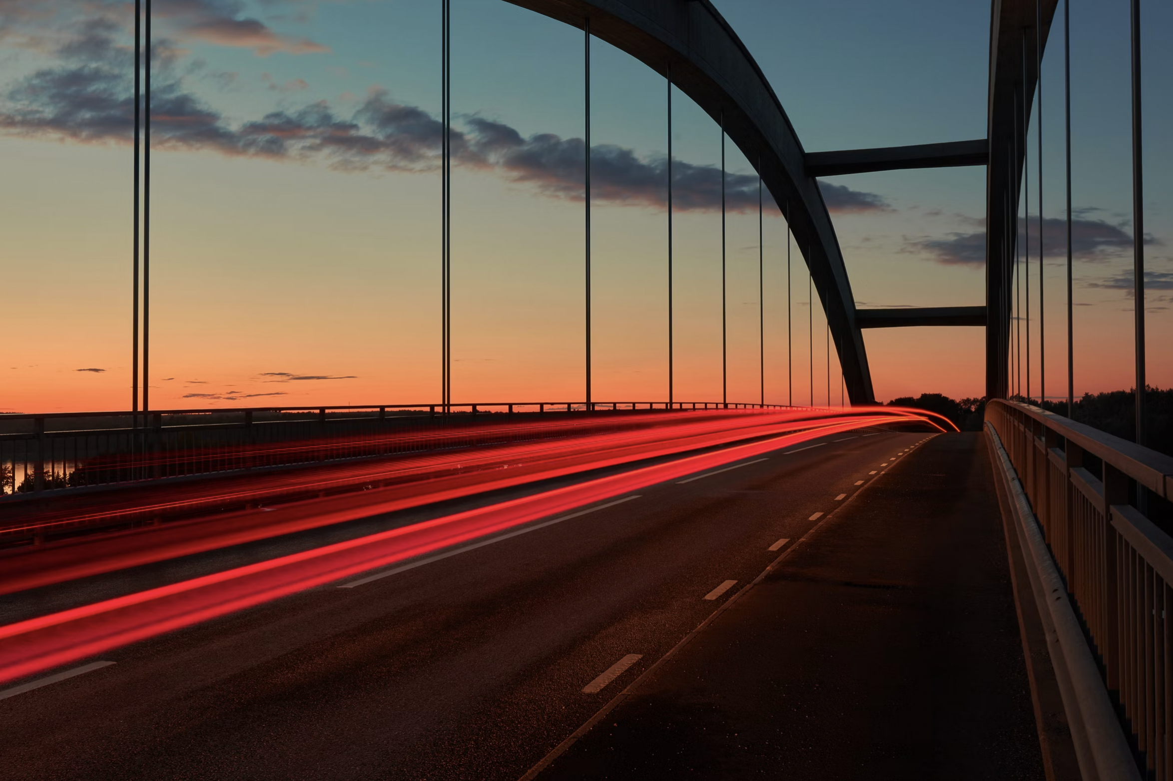 Sunset over a modern suspension bridge with red car light trails on the roadway.
