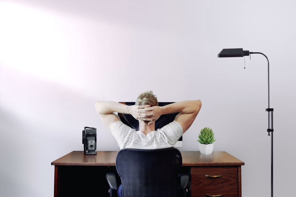 Person seen from behind, arms folded behind head while seated at a desk in an office setup with a vintage camera, a lamp, and two small plants on the desk.