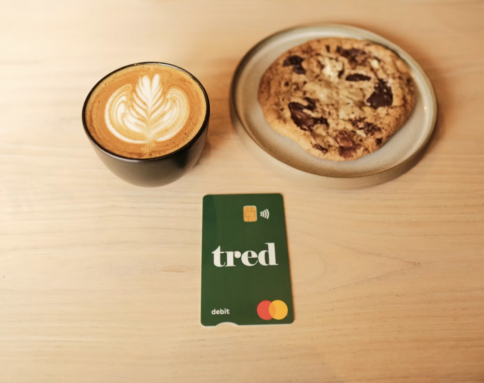 Latte with leaf latte art and a chocolate chip cookie on a wooden table, with a green debit card labeled 'tred' in front.