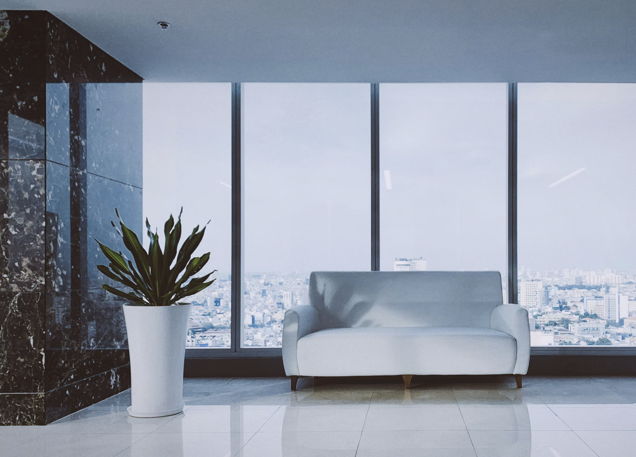 Modern office lobby with a white sofa, tall marble wall, and a potted plant, overlooking a city skyline through large windows.