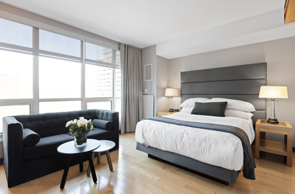 Hotel bedroom with a large window, gray headboard bed, nightstands, lamps, and a dark blue sofa by the seating area.