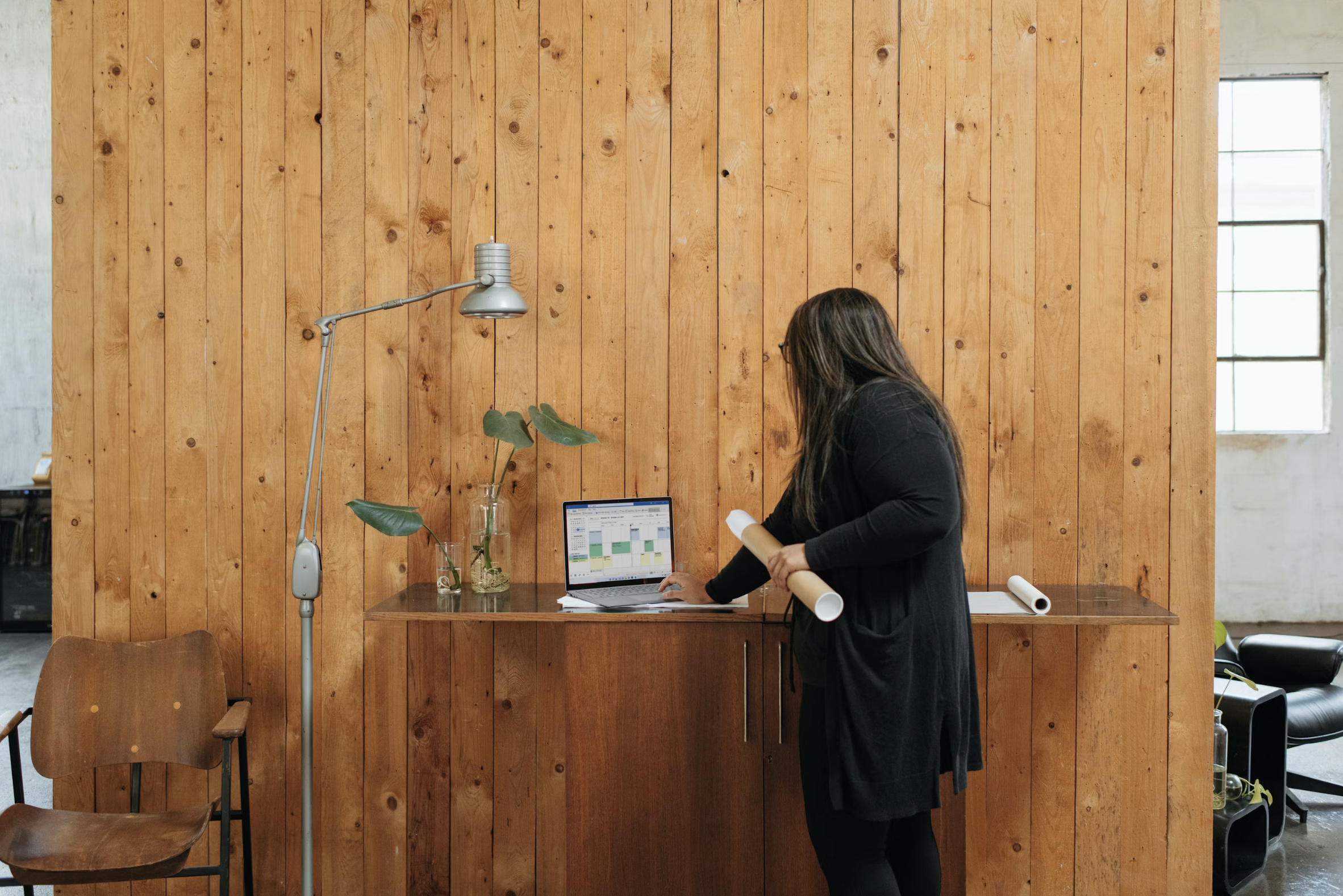 Woman standing at a wooden desk with a laptop open, holding a rolled paper in a modern office with a wood-paneled wall and a tall adjustable lamp nearby.