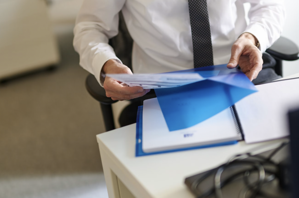 Person in a white shirt and patterned tie sorting or organizing blue folders on a desk, with documents in hand.