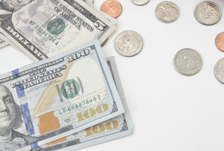 Stack of US currency including $100 bills and scattered coins on a white surface, money ready for counting or exchange.