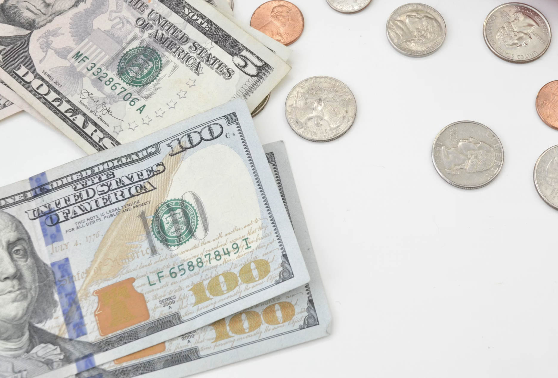 Stack of US currency including $100 bills and scattered coins on a white surface, money ready for counting or exchange.