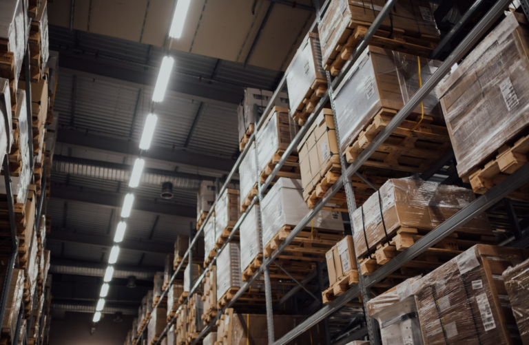 Industrial warehouse with tall steel racks and stacked pallets on both sides, illuminated by bright overhead lights in a high-ceiling storage area.