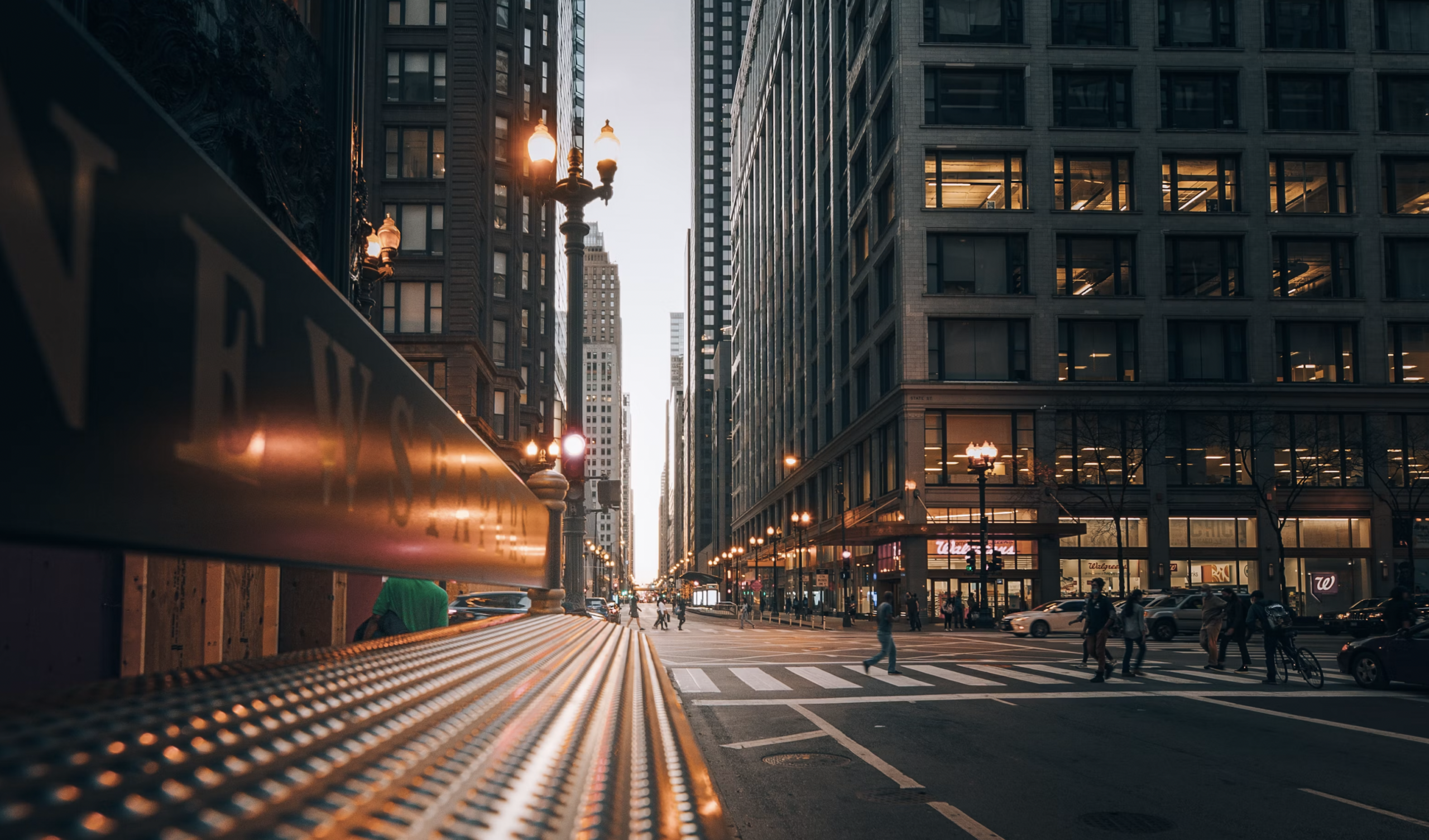 City street scene at dusk with tall buildings, glowing streetlights, and pedestrians crossing a crosswalk near a bus shelter railing in the foreground