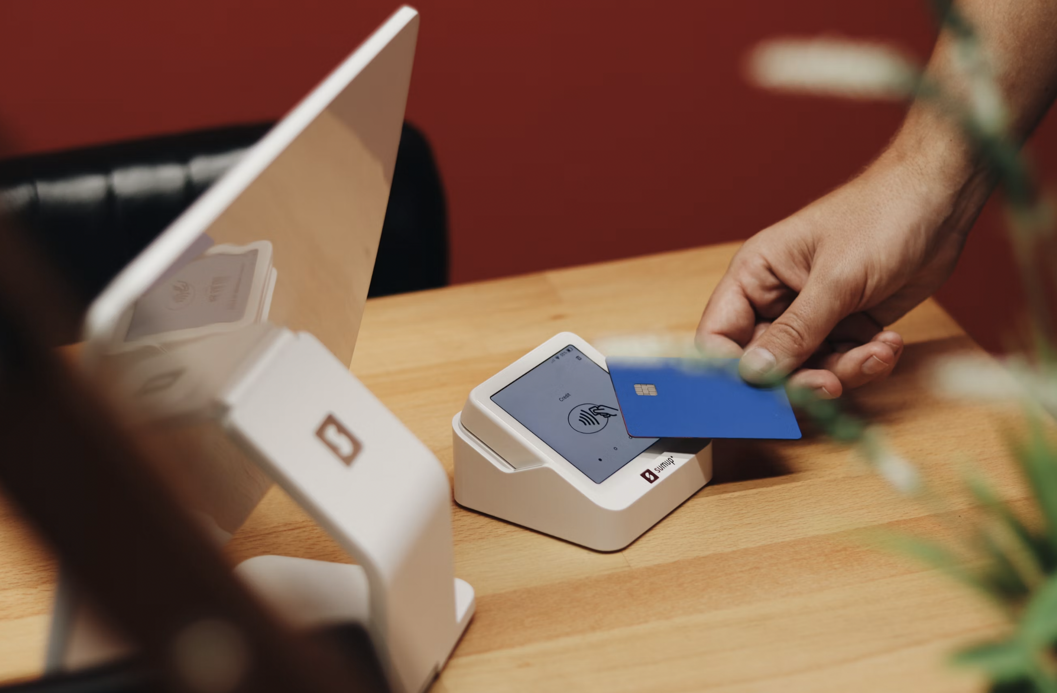 Close-up of a hand holding a blue card near a white payment terminal on a wooden checkout counter, ready to pay.