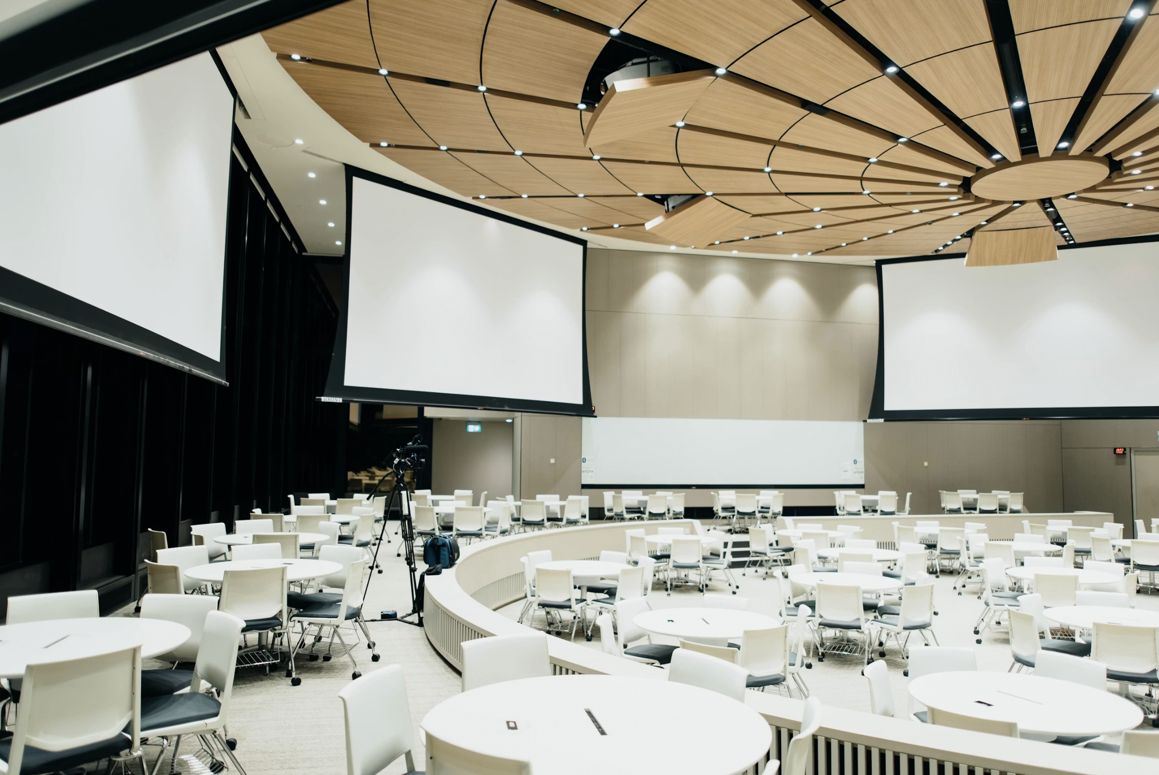 Conference hall with a circular wooden ceiling, white tables and chairs arranged for a presentation, and two large projection screens on opposite walls.