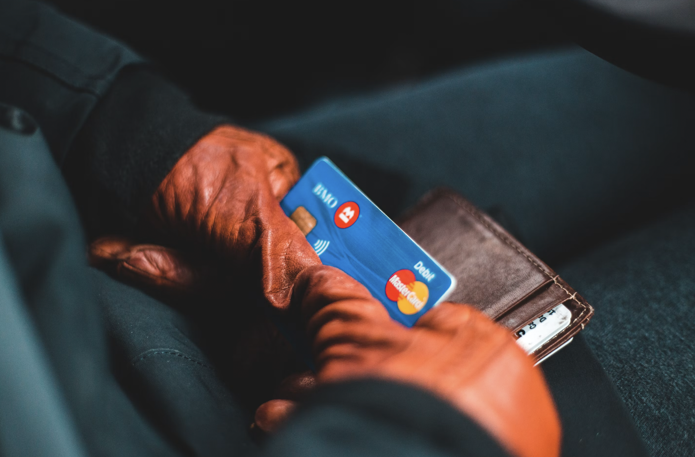 Person holding a blue credit card near a brown wallet on their lap, dark background.