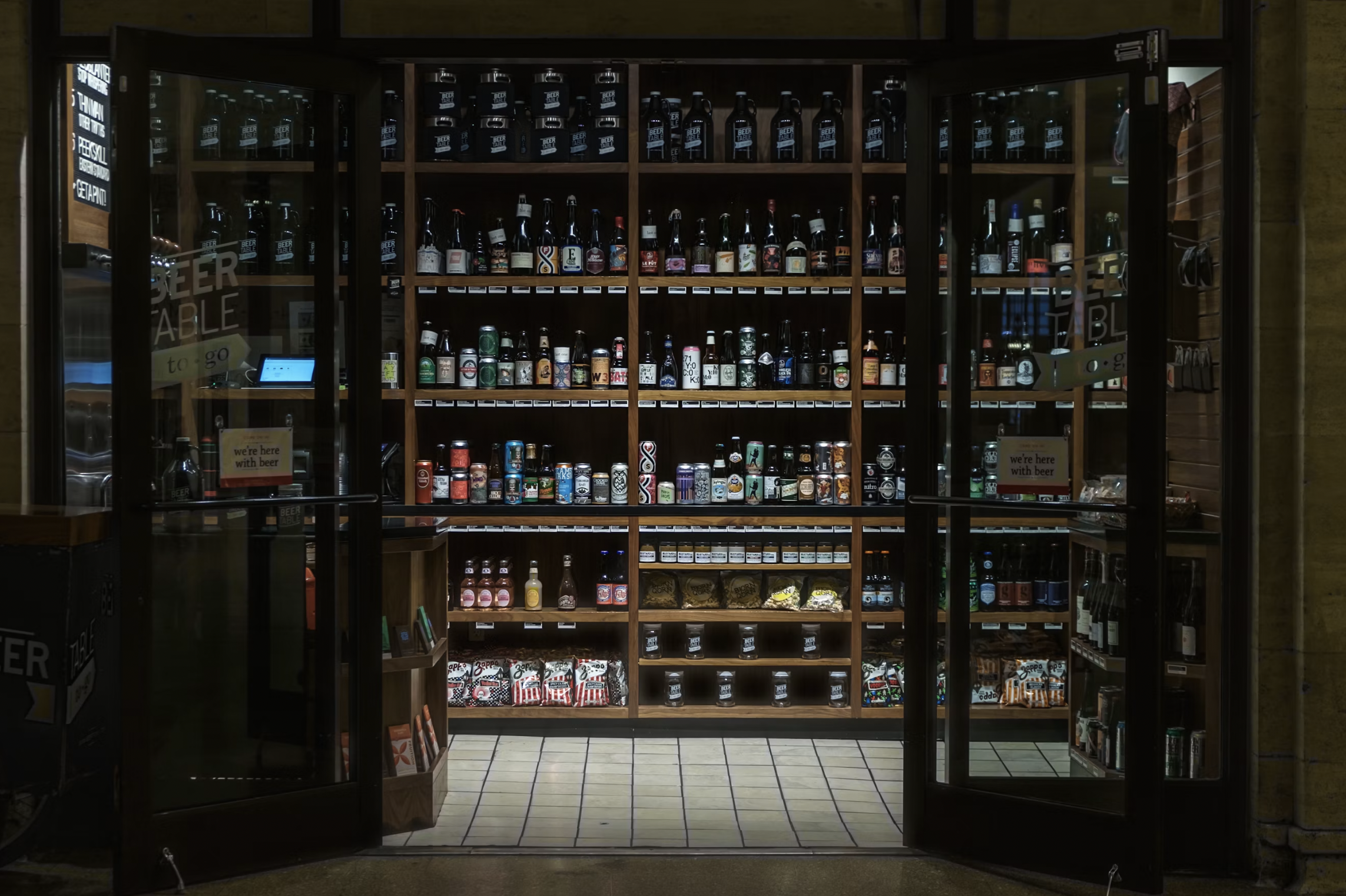 Glass-front beer display with multiple shelves of bottles and cans in a store refrigerator, labeled shelves and a wooden interior.